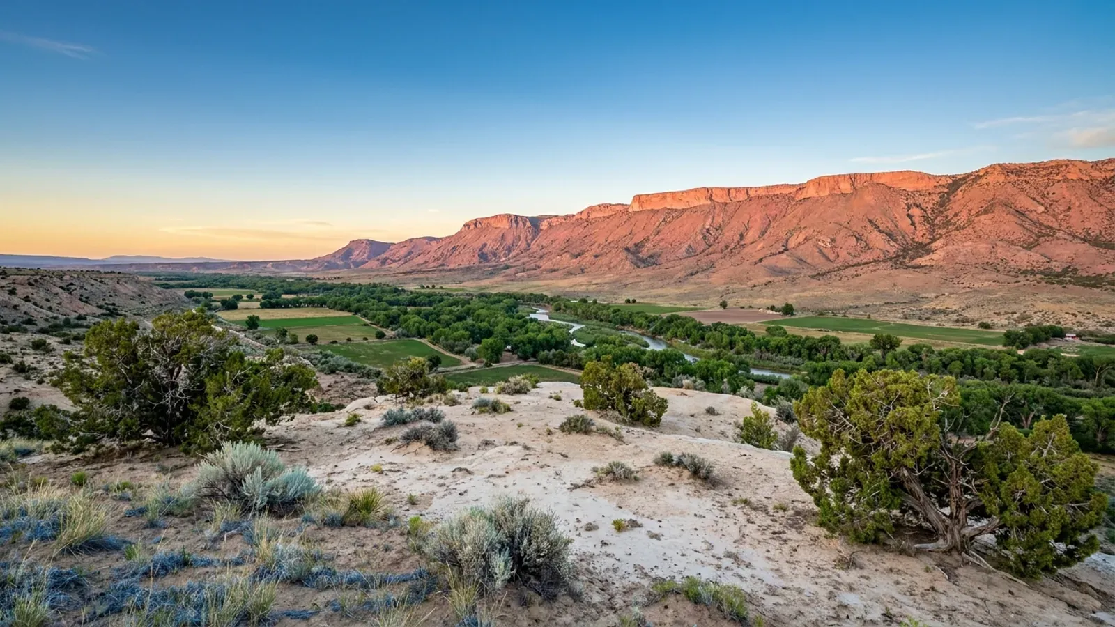 High desert mesa above the Rio Grande valley, a line of cottonwoods along the river and a long mountain range glowing pink at sunset beyond.