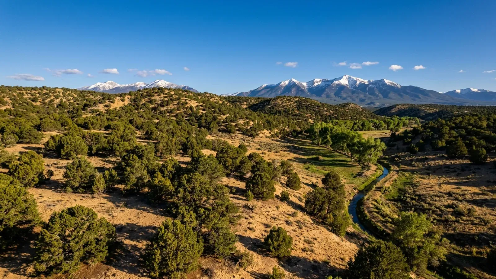 Piñon-juniper hills falling to a green stream valley, below high mountains with snow on the peaks in northern New Mexico.